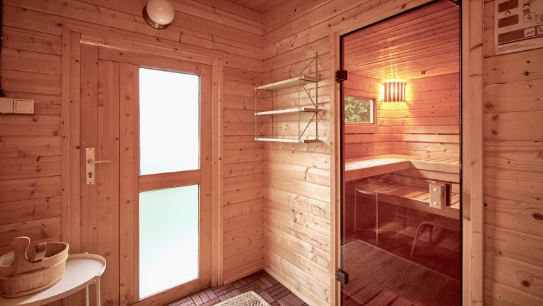 Innenansicht einer Sauna mit Holzwänden, einer Glastür und einer Bank. Ein Holzeimer steht auf einem Tisch.