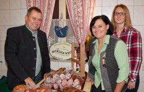 Three people are standing in front of a table with sausage products in a farm store. In the background is a sign with the inscription 'Bucklige Welt'.