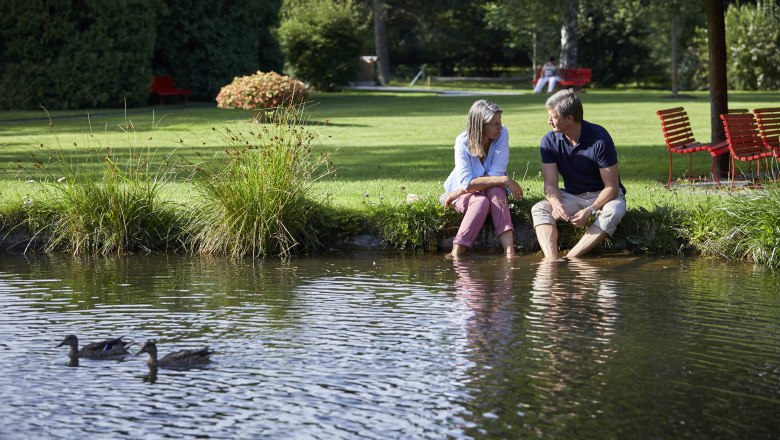 Ein Paar sitzt am Ufer eines Teichs im Kurpark Bad Schönau, die Füße im Wasser. Zwei Enten schwimmen im Vordergrund.