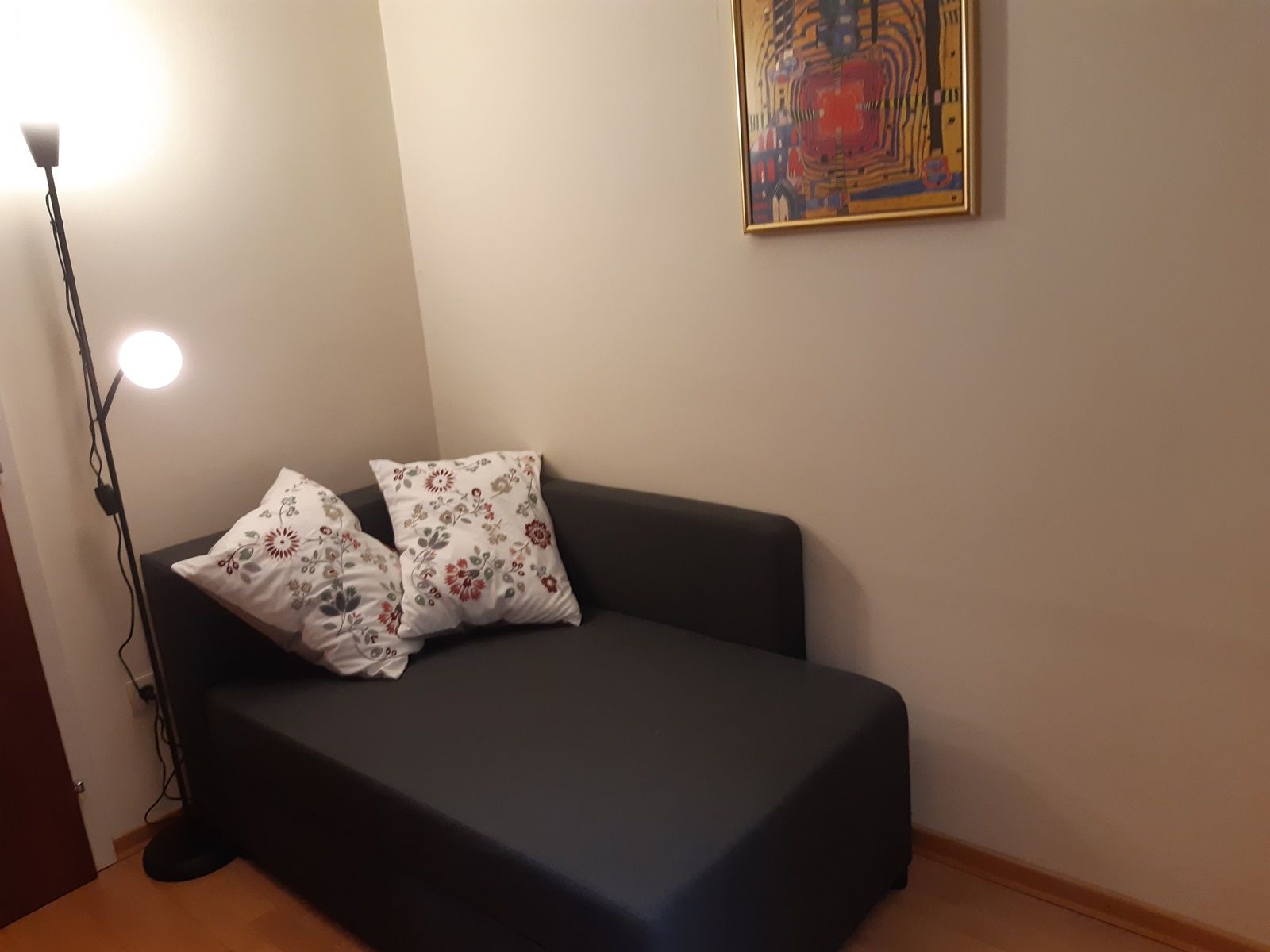 Gemütliche Leseecke mit grauem Sofa, zwei gemusterten Kissen, Stehlampe und abstraktem Bild an der Wand.