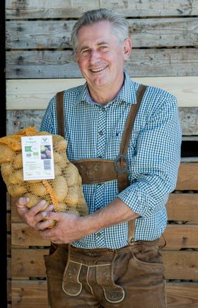 Zwei Männer vor einer Holzwand, einer hält Kartoffeln, der andere einen Korb mit Kürbissen.