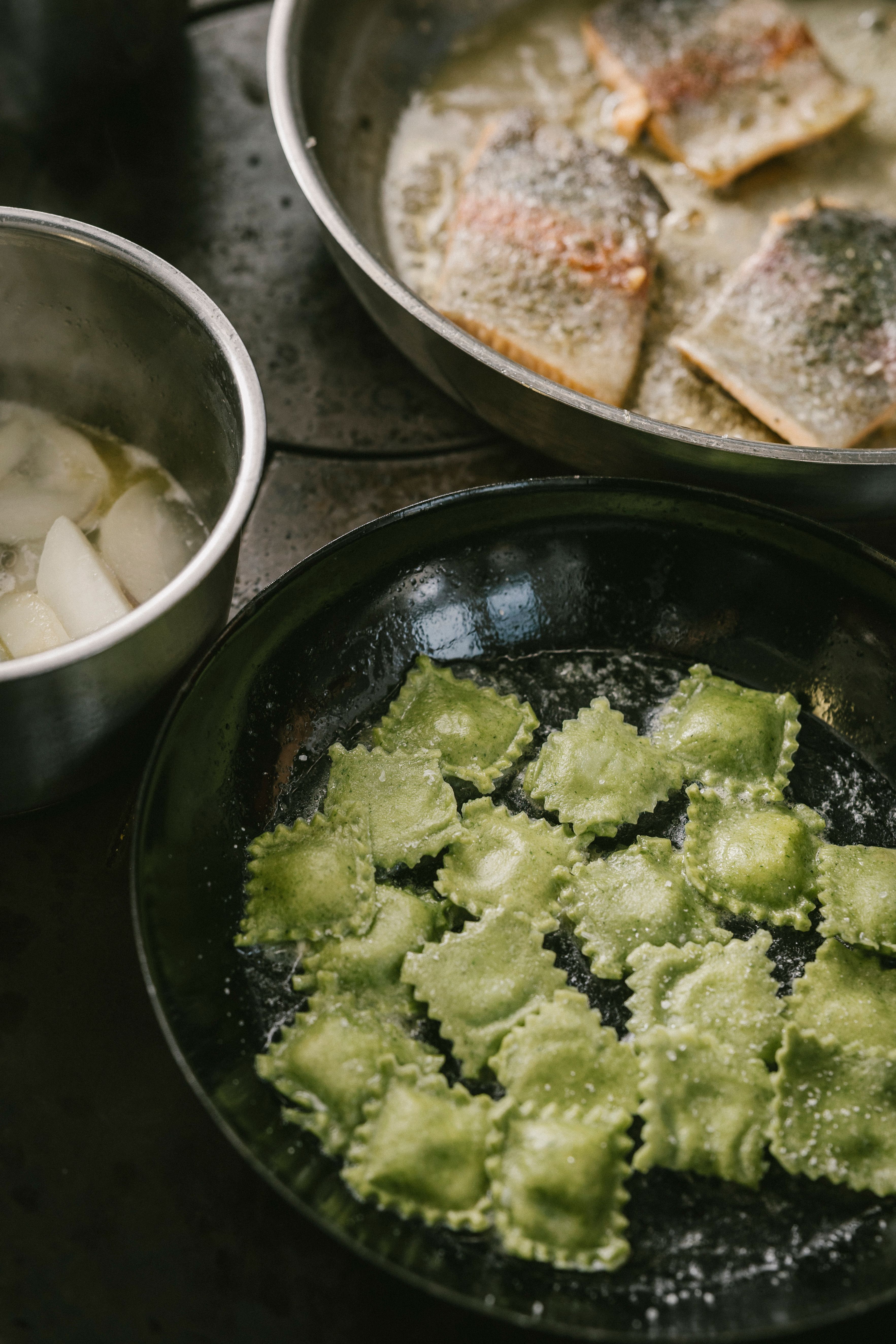 Mehrere Edelstahlschüsseln mit Fischfilets, Kartoffelstücken und grünen Ravioli in einer Küche.