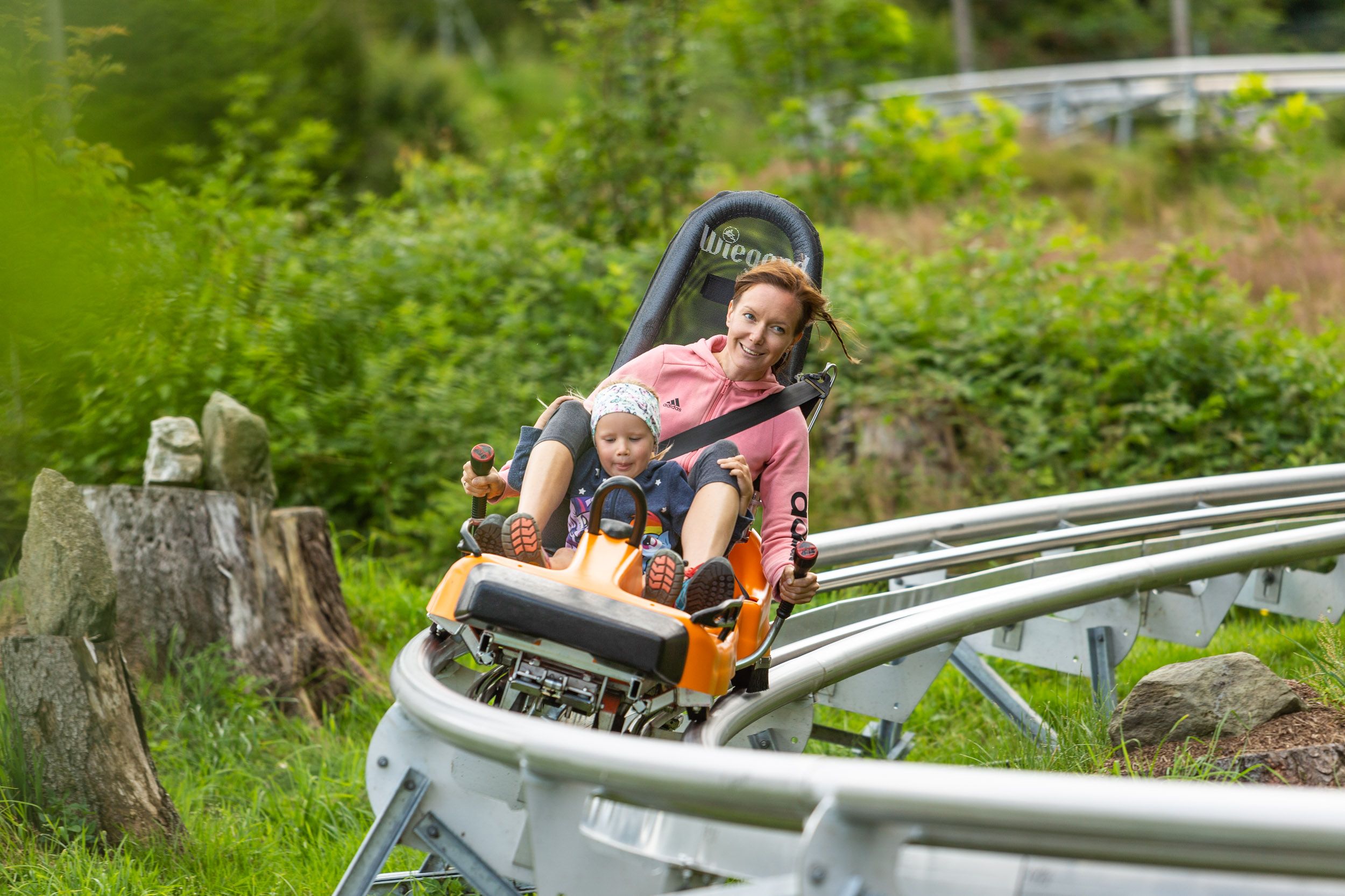 Eine Frau und ein Kind fahren auf einer Sommerrodelbahn durch eine grüne Landschaft.