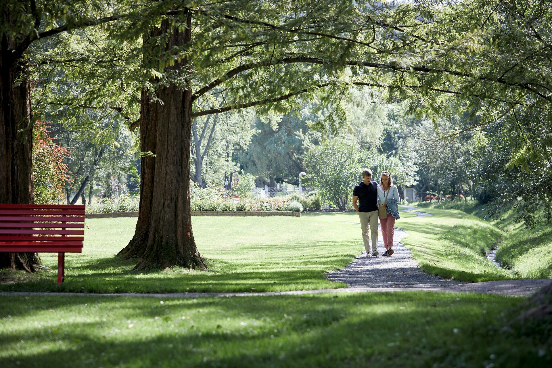 Eine Frau und ein Mann halten sich bei der Hand und spazieren durch einen Park, indem rote Sitzbänke seitlich in der grünen Wiese stehen.