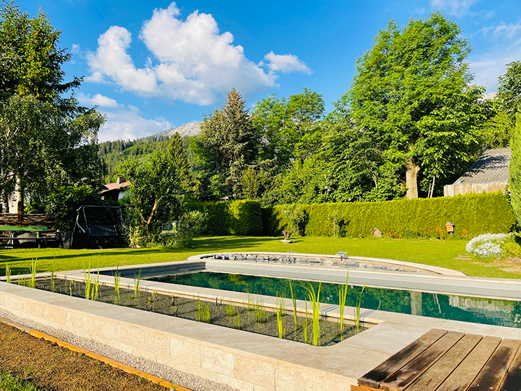 A well-tended garden with a rectangular pool, surrounded by trees and hedges, under a blue sky with white clouds.