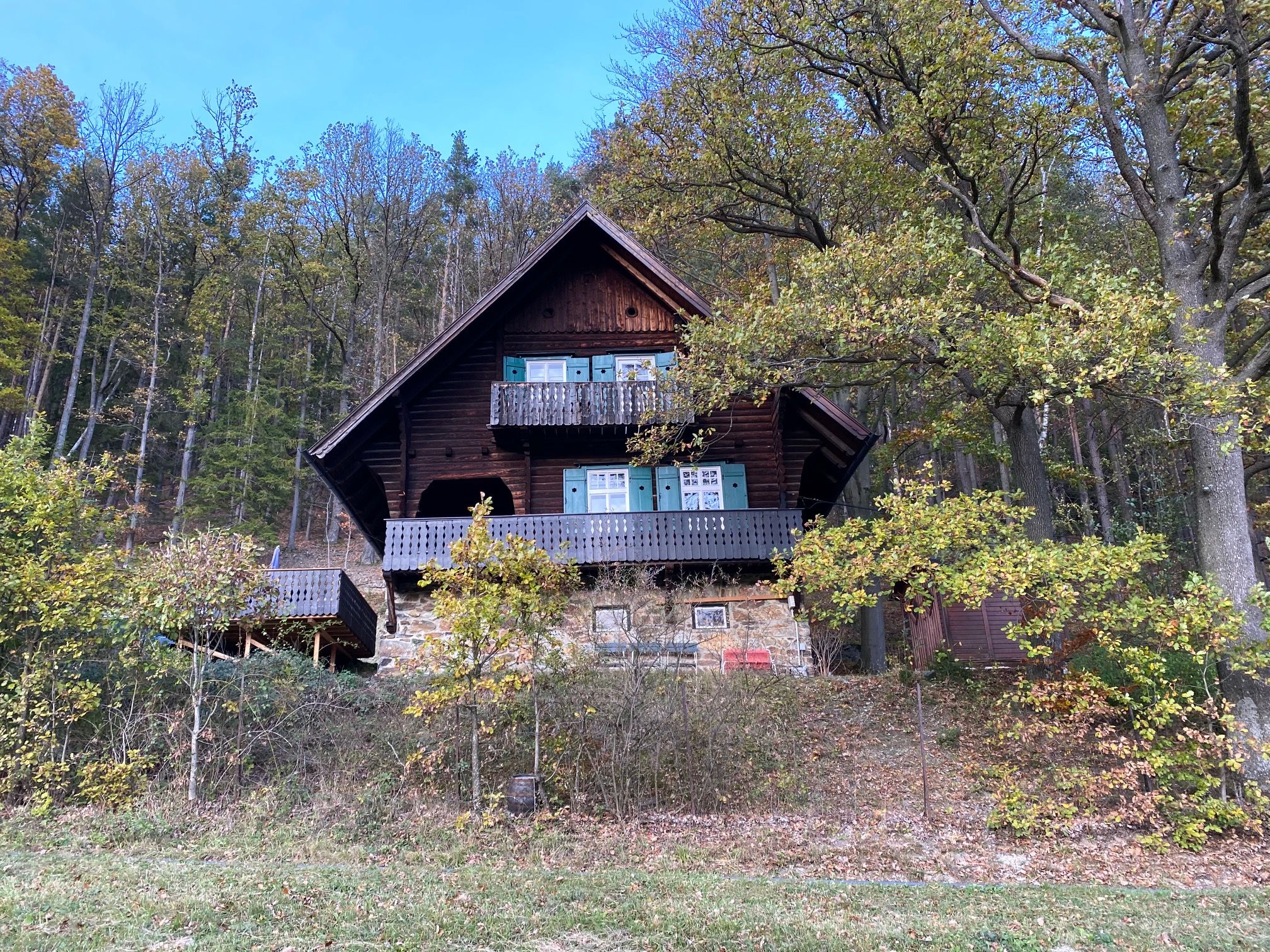 Ein rustikales Holzhaus mit Balkon steht am Waldrand, umgeben von Bäumen und herbstlichem Laub.