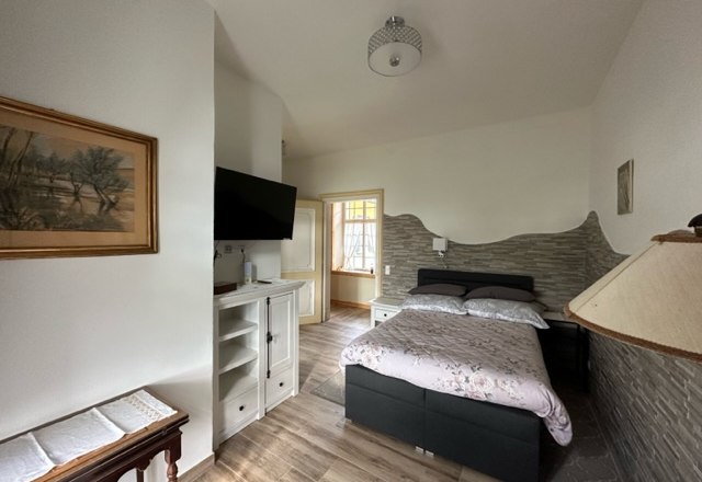 Ein gemütliches Zimmer mit einem Einzelbett, einem Fernseher an der Wand und einem kleinen Tisch. Helle Wände und Holzboden schaffen eine einladende Atmosphäre.