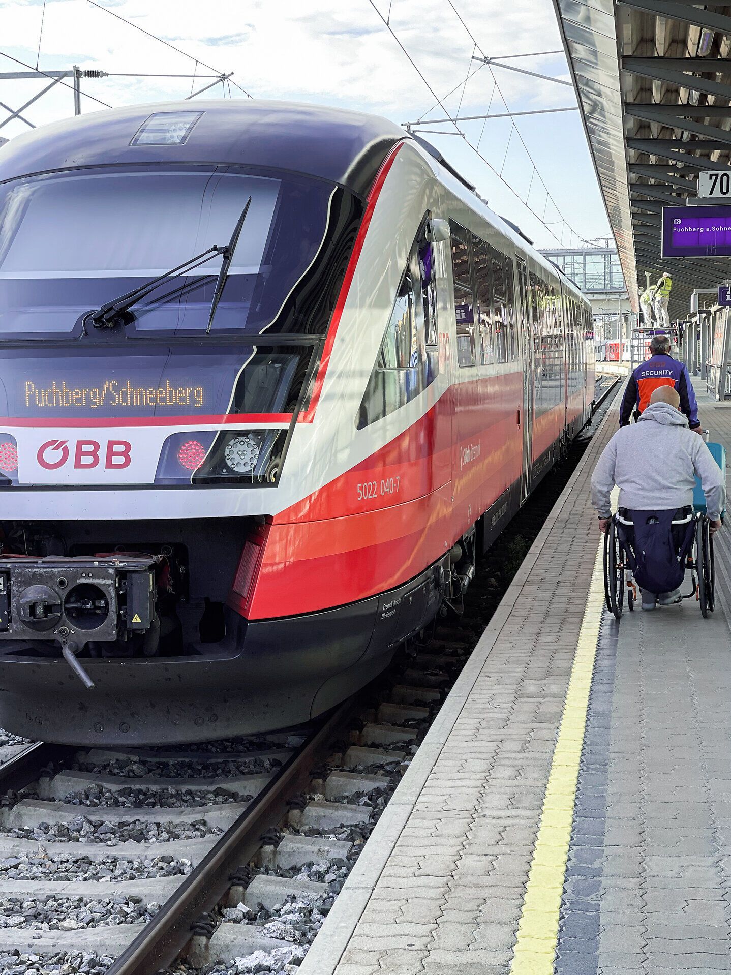 Am Bahnhof der Wiener Alpen wartet ein moderner Zug auf die Reisenden, während ein Passagier im Rollstuhl geduldig auf seine Abfahrt vorbereitet. Die Atmosphäre ist geprägt von Vorfreude und der Möglichkeit, die atemberaubenden Landschaften der Region zu erkunden. Hier beginnt das Abenteuer in den malerischen Bergen.