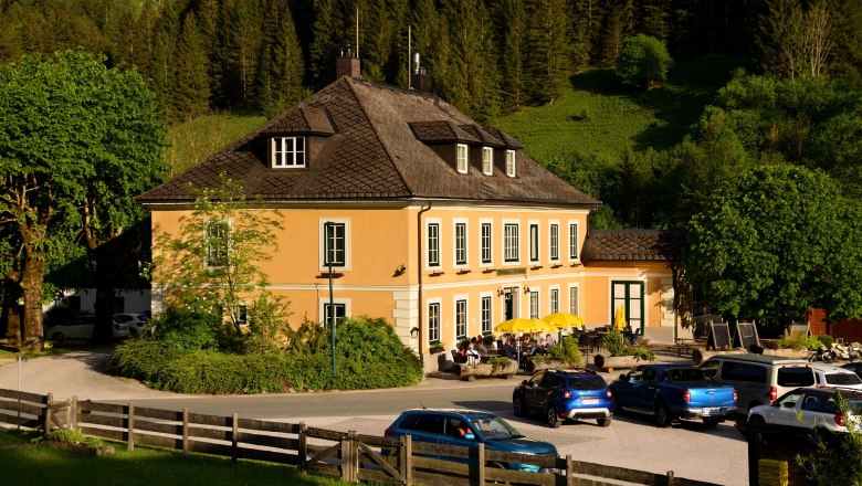 A yellow pub with a terrace and parasols, surrounded by trees and cars, in front of a wooded hill.
