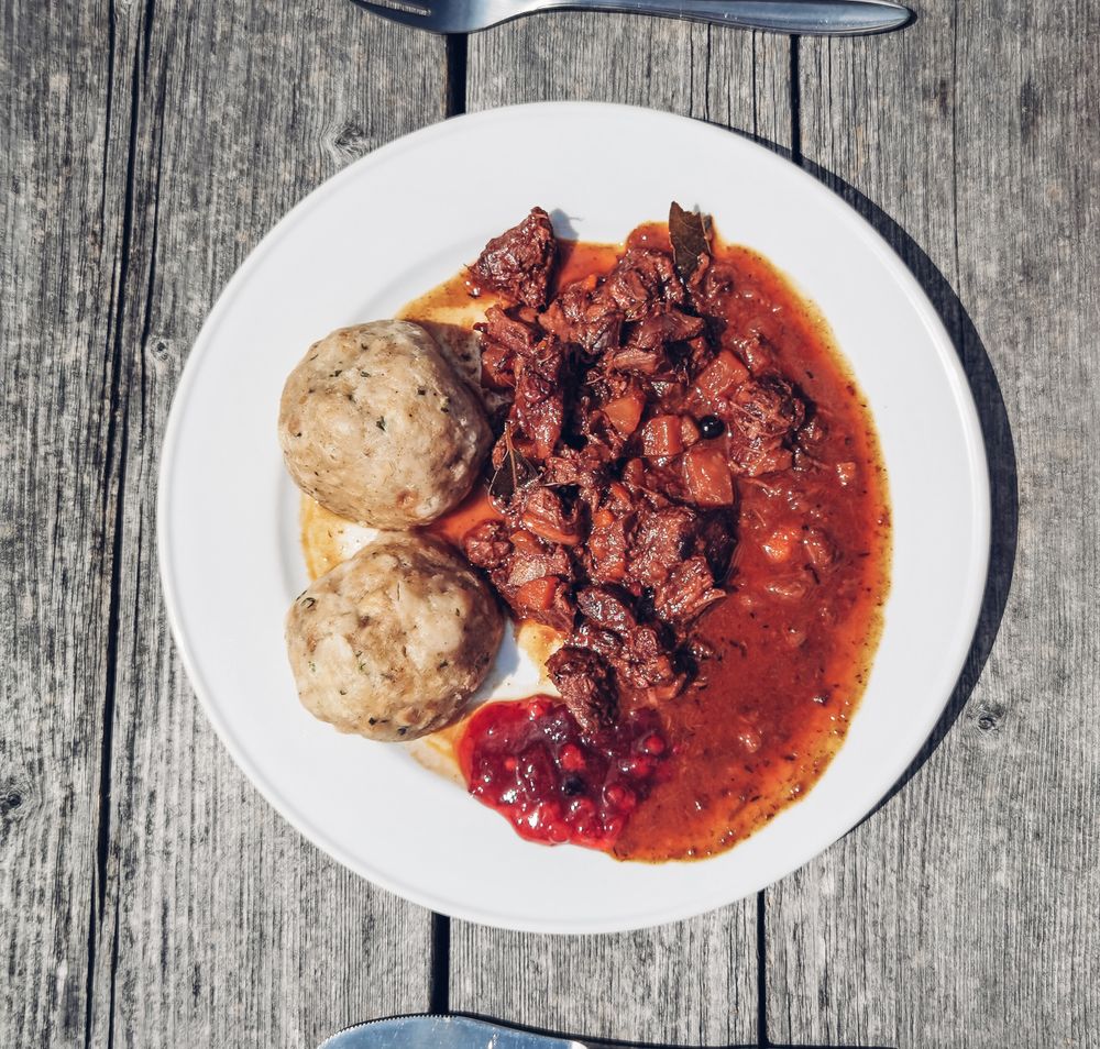 Die sanften Hügel und die goldene Abendsonne schaffen eine magische Atmosphäre, während Wanderer den Pfad entlang der Wiesen erkunden. Die beeindruckende Aussicht auf die umliegenden Berge lädt dazu ein, innezuhalten und die Schönheit der Natur zu genießen.