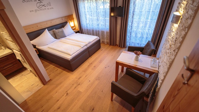 Gem&uuml;tliches Doppelzimmer mit Holzboden, Doppelbett, kleinem Tisch und St&uuml;hlen, dekorativen Vorh&auml;ngen und einem Fernseher an der Wand.