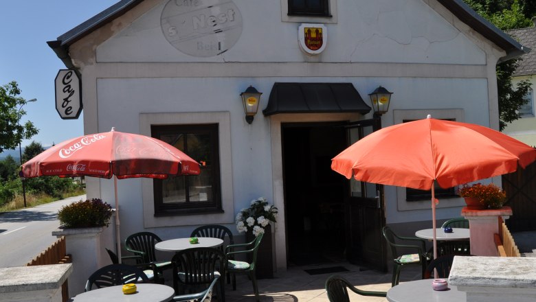 Außenansicht eines Cafés mit Terrasse, Tischen und Sonnenschirmen.
