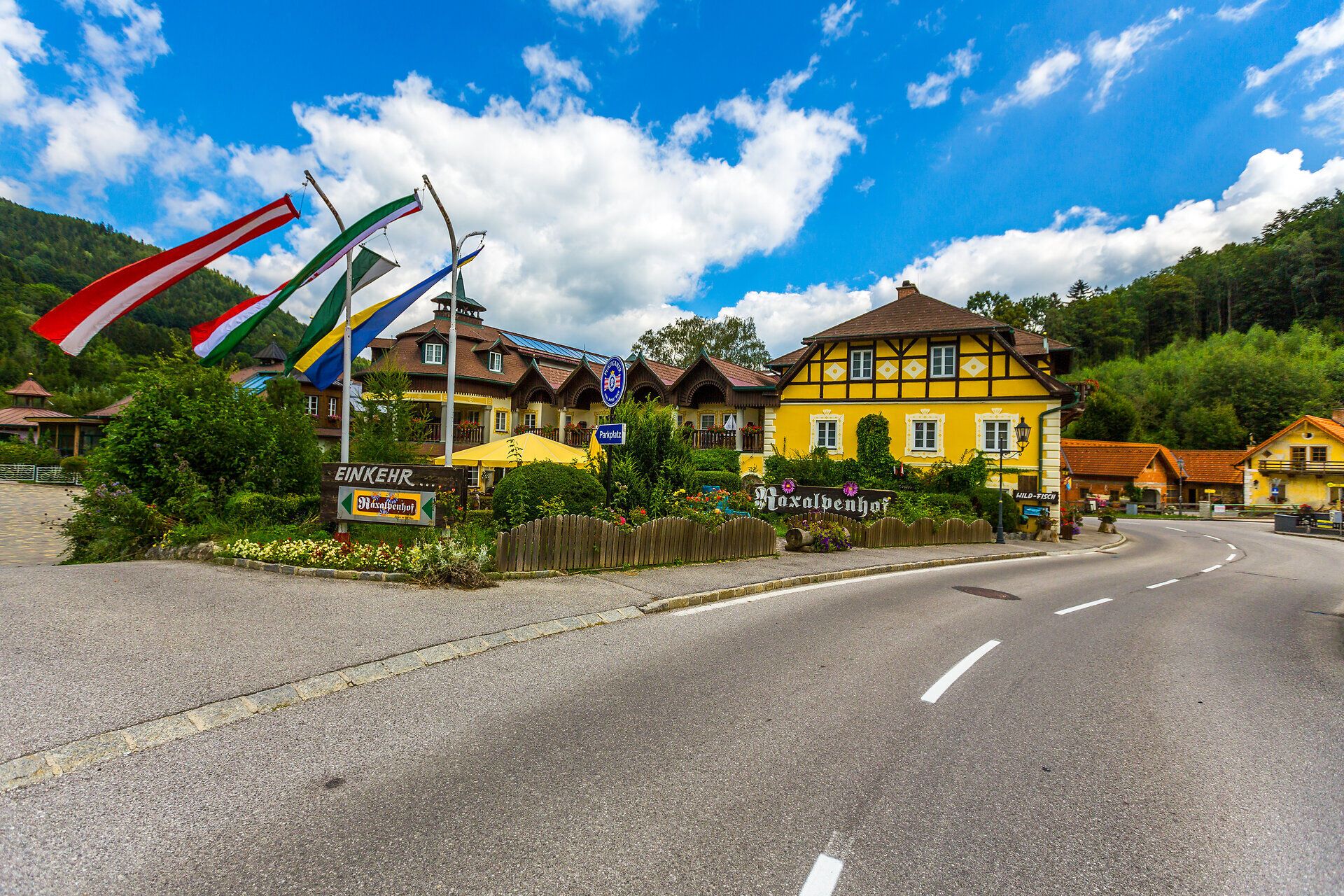 Raxalpenhof liegt direkt an der Ortsstraße, vor dem Haus steht ein blauer Brunnen, Blumen und Sträucher säumen die Balkone und en gesamten Vorderbereich des Hauses