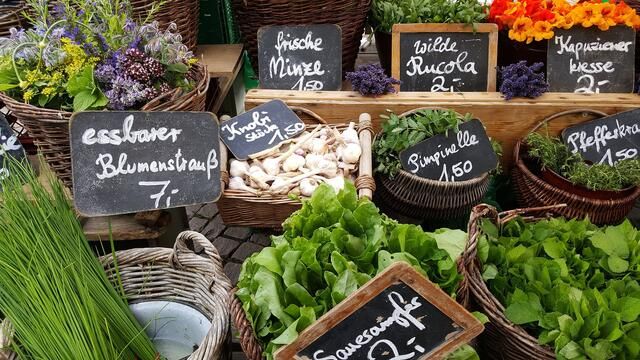 Marktstand mit Kräutern und Gemüse in Körben, beschriftet mit Tafeln.