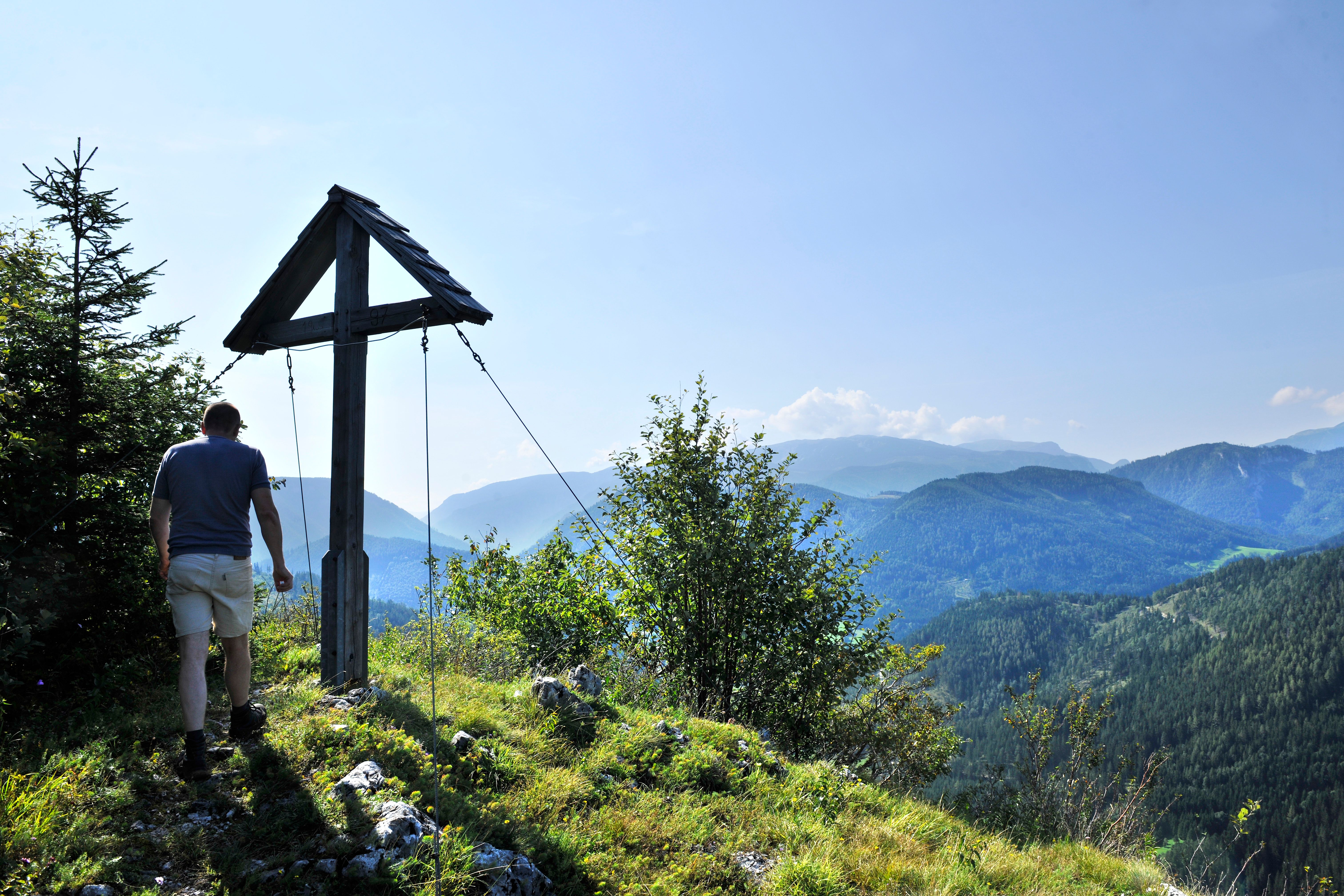 Ein Wanderer steht neben einem Gipfelkreuz mit Blick auf bewaldete Berge.