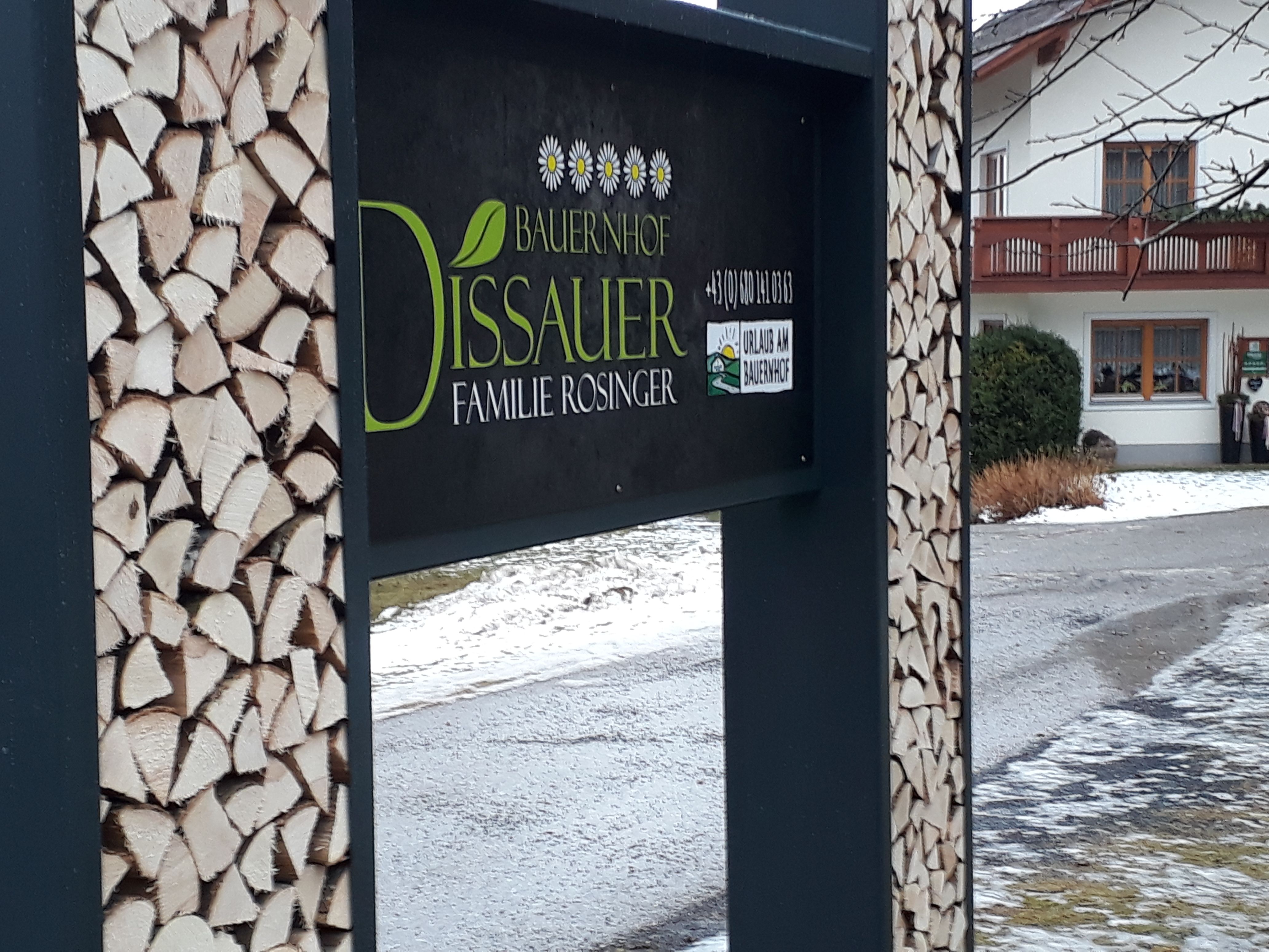 Schild eines Bauernhofs mit Holzstapeln und einem Haus im Hintergrund.
