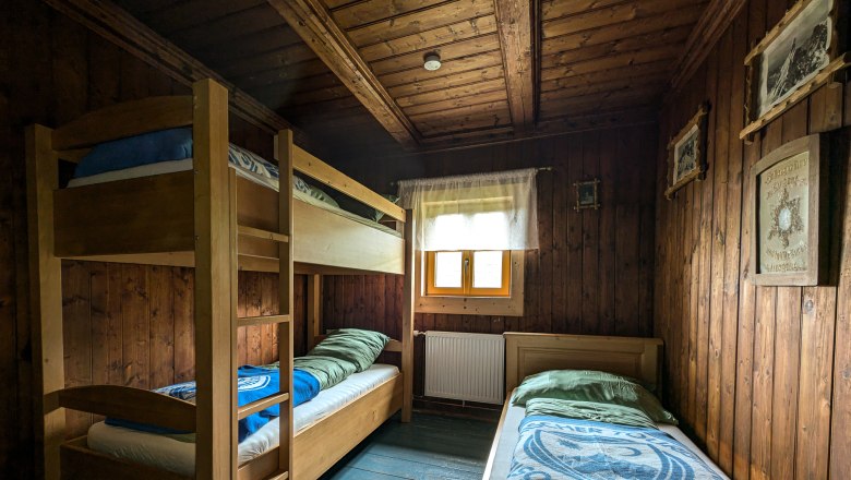 Wood-paneled triple room with bunk bed and single bed, window with curtain, rustic furnishings.