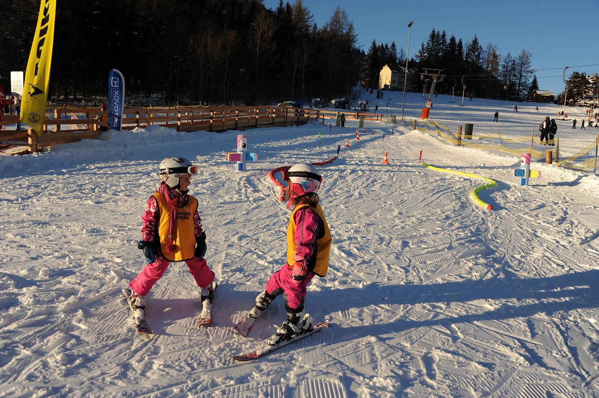 Zwei Kinder auf kleinen Schi stehen auf einer kurzen flachen Piste auf der Hütchen aufgestellt sind.