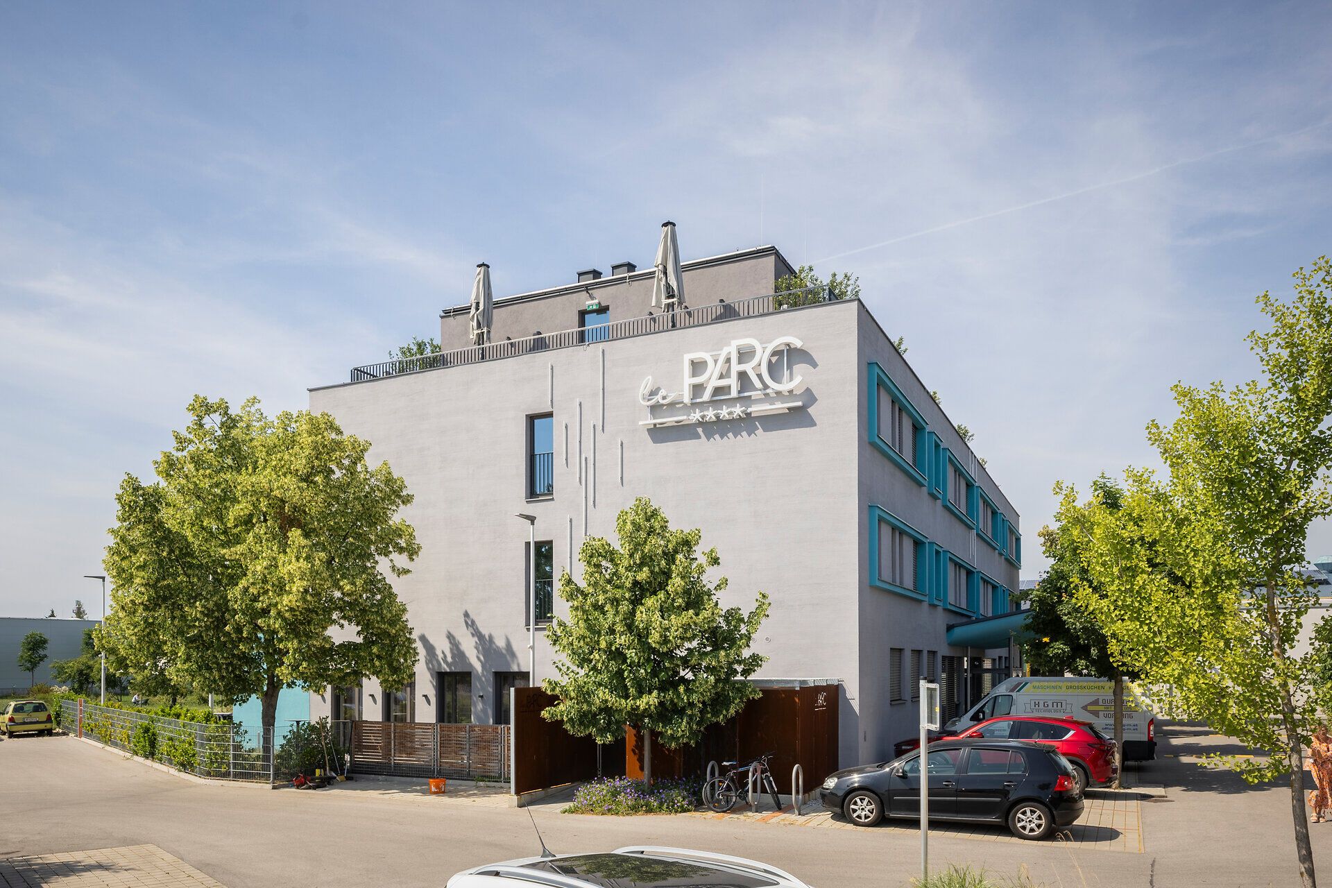 Vor dem Hotel leParc mit Dachterrasse und großer "le PARC" Aufschrift parken Autos und ein Rad, zwischendurch stehen einige Bäume.