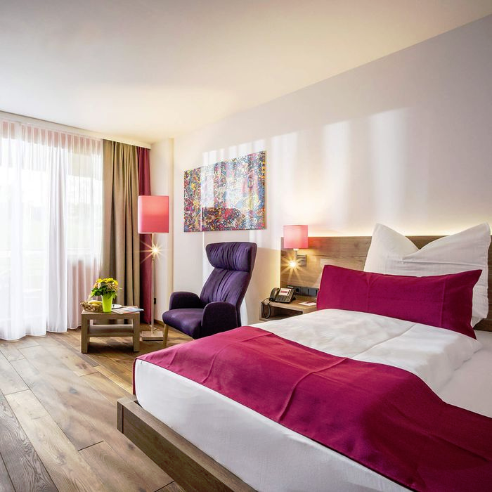 Gemütliches Zimmer mit Doppelbett, Ohrensessel, Tisch, Lampen, einem Wandbild und Holzboden.