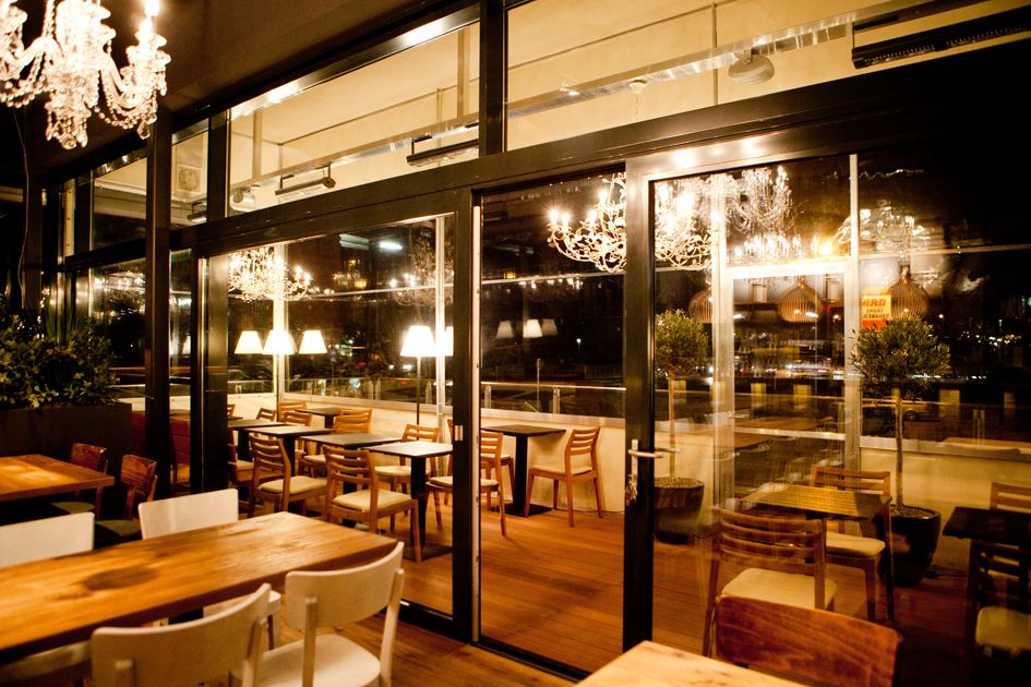 Ein stilvoll eingerichtetes Restaurant mit Holztischen und Stühlen, beleuchtet von Kronleuchtern und Tischlampen, mit Blick auf eine Terrasse.