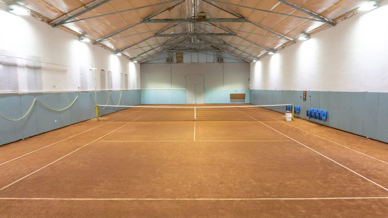 Innenansicht eines leeren Tennisplatzes in einer Halle mit braunem Bodenbelag und blauen Wänden.
