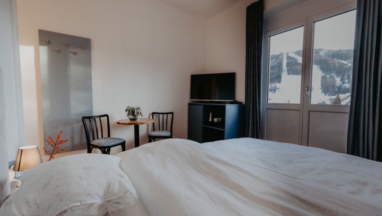 A cozy hotel room with a bed, two chairs, a table and a TV. You can see a snow-covered ski slope through the window.