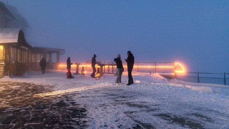 Menschen stehen auf verschneiter Bergterrasse im Nebel, beleuchtet von warmen Lichtern bei D&auml;mmerung.