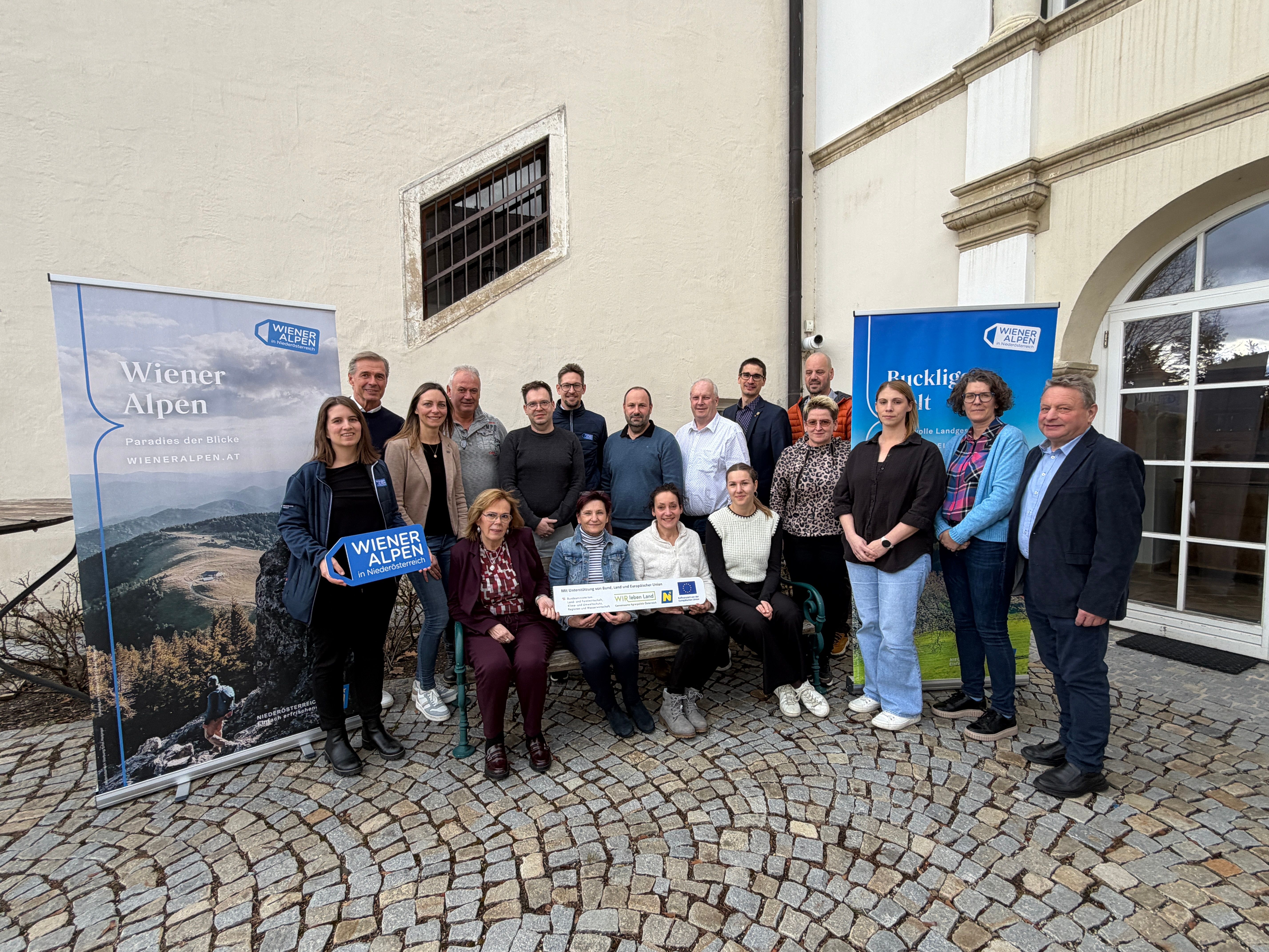 Mehrere Personen stehen in einem Innenhof und halten ein Wiener Alpen Schild und ein LEADER-Schild in den Händen.