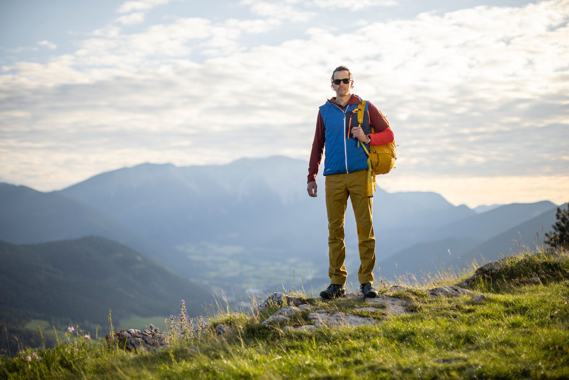 Eine Person mit Rucksack steht auf einem grasbewachsenen Hügel vor einer weiten Berglandschaft