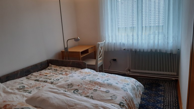 Ein Zimmer mit einer ausgezogenen Couch als Bett, einem Schreibtisch mit Stuhl und einem Fenster mit Vorh&auml;ngen.