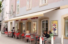 Außenansicht des Cafés mit roten Stühlen und Gästen an Tischen.