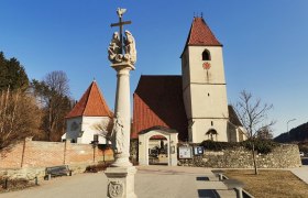 Pfarrkirche Unter-Aspang mit Säule im Vordergrund und blauem Himmel.