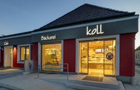 Exterior view of the bakery in the evening with illuminated sign 'Bäckerei Koll' and 'Cafe'.