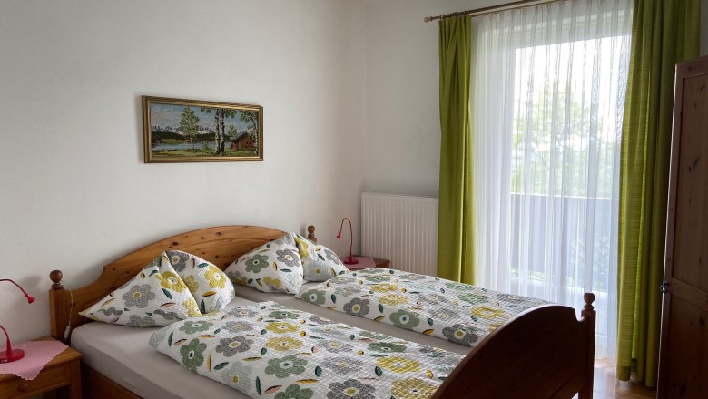 Ein gemütliches Doppelzimmer mit Holzmöbeln, einem großen Fenster mit grünen Vorhängen und einem Landschaftsbild an der Wand.