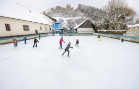 Menschen eislaufend auf einem Platz in Kirchschlag, im Hintergrund auf einem Hügel eine Burgruine.
