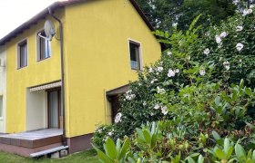 Gelbes Haus mit Terrasse und bl&uuml;henden Str&auml;uchern im Vordergrund.