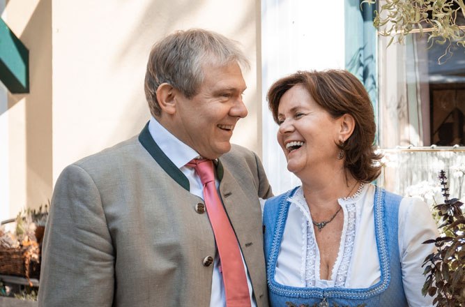 Egy férfi és egy nő hagyományos bajor ruhában nevetnek együtt egy épület előtt.