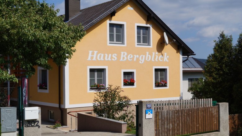 Gelbes Haus mit der Aufschrift 'Haus Bergblick', umgeben von Bäumen und einem Holzzaun.