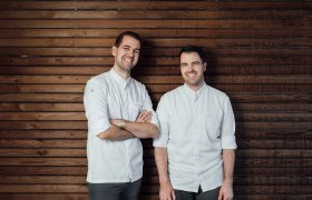 Zwei Männer in weißen Hemden stehen vor einer Holzwand und lächeln.