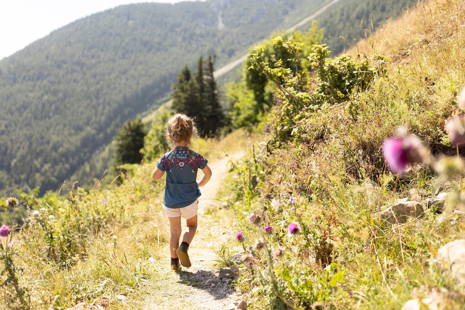 Ein fröhliches Kind erkundet den malerischen Wanderweg, umgeben von üppigem Grün und bunten Wildblumen. Die sanften Hügel und majestätischen Berge im Hintergrund laden zu unvergesslichen Abenteuern in der Natur ein. Hier, wo die frische Bergluft die Sinne belebt, wird jeder Schritt zu einem Erlebnis.