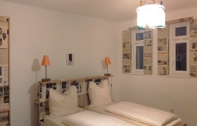 Hotelzimmer mit Doppelbett, Zeitungsdekoration an Wänden und Fenstern, zwei orangefarbene Lampen.
