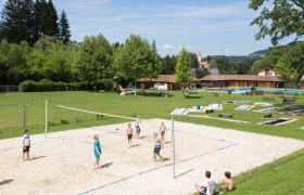 Kinder spielen Volleyball auf einem Sandplatz im Freibad Aspang, umgeben von grüner Wiese und Bäumen.