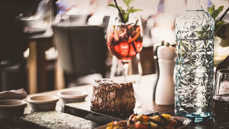 Ein saftiges Steak auf einem heißen Stein serviert, begleitet von Gemüse, einer Karaffe Wasser und einem Cocktailglas.