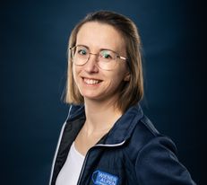 Portraitfoto Wiener Alpen Mitarbeiterin, die eine Jacke mit Wiener Alpen Logo trägt und vor einem dunklem Hintergrund steht.