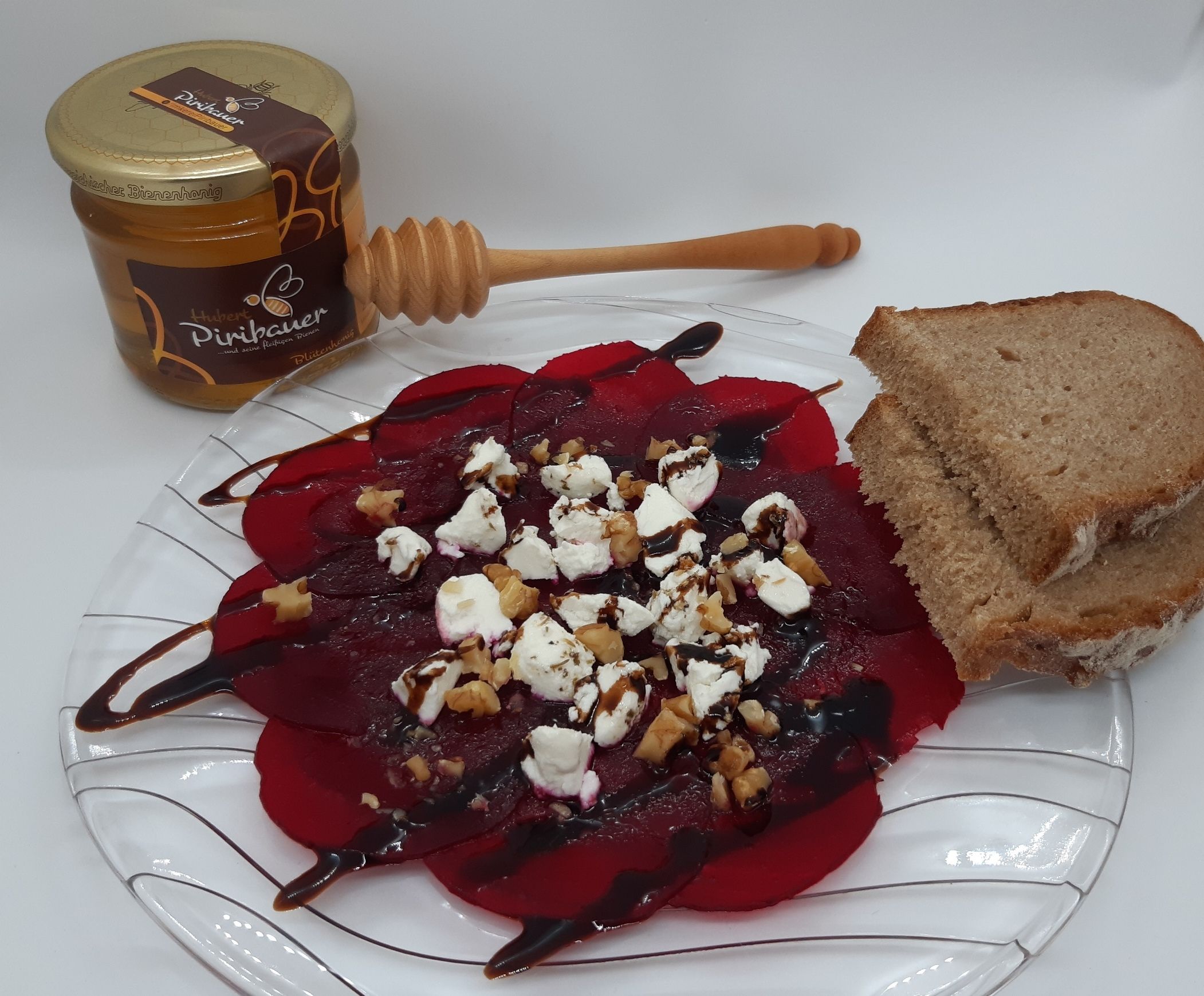 Rote Rüben Carpaccio mit Ziegenfrischkäse, Nüssen, Balsamico und Brot auf Glasteller, daneben Honig.