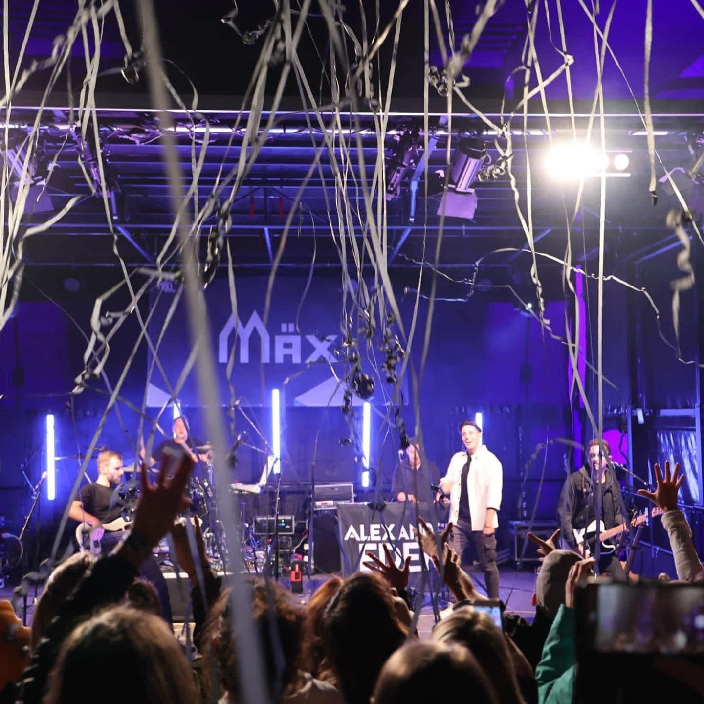 Livekonzert auf einer beleuchteten Bühne mit Band, herabfallendem Konfetti und jubelndem Publikum im Vordergrund.