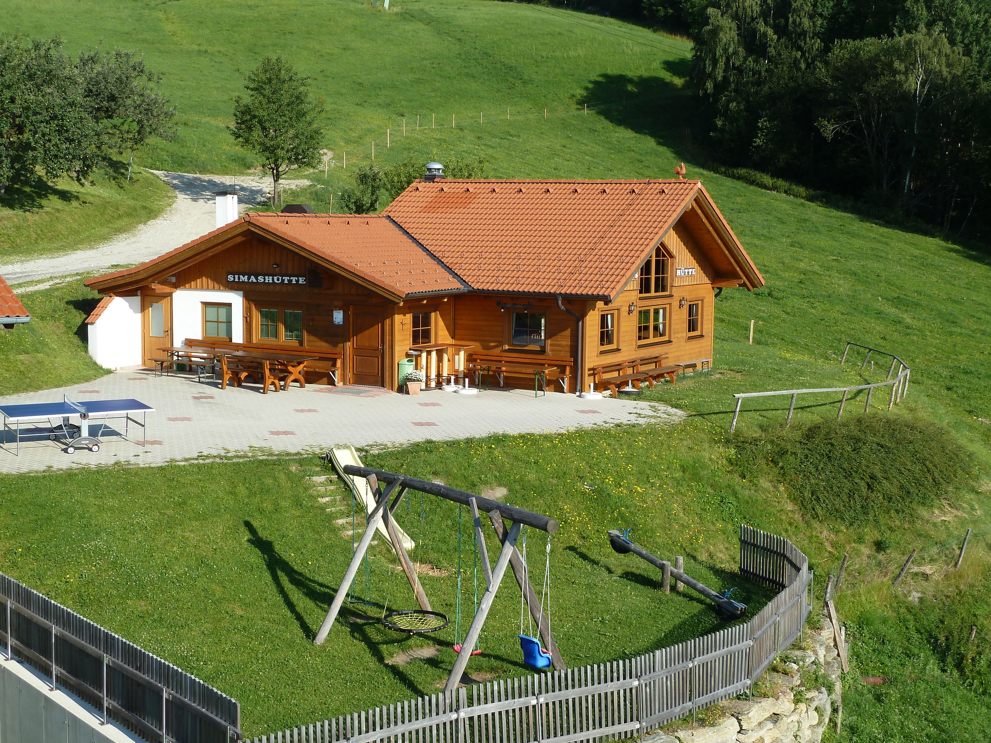 Eine Berghütte namens Simashütte mit rotem Dach, umgeben von grünen Wiesen und einem Spielplatz mit Schaukel.