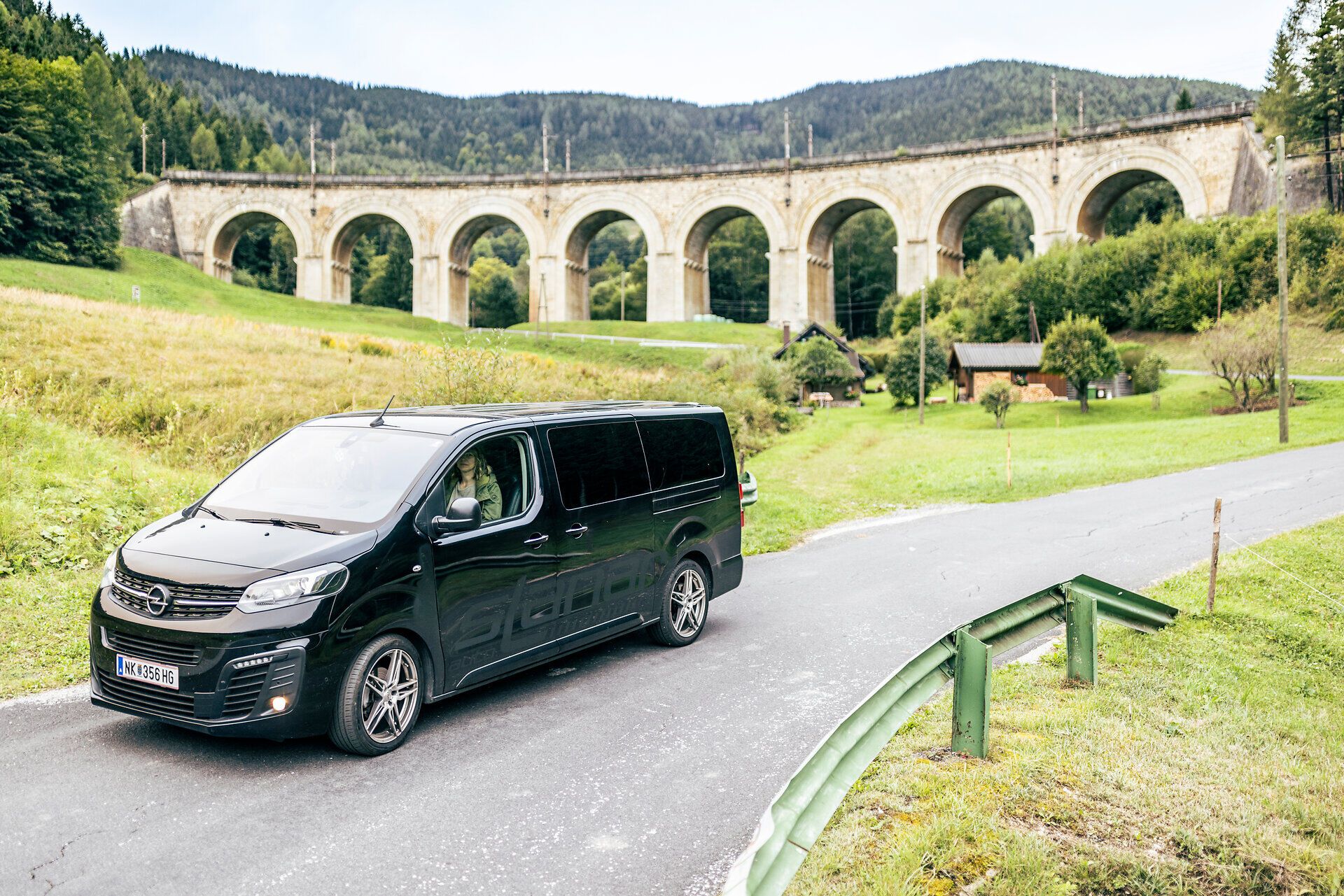 Ein schwarzer Kleinbus, der RUF Bus, holt gerade eine Frau in einem schönen orangen Kleid und einen Wanderer ab. 
