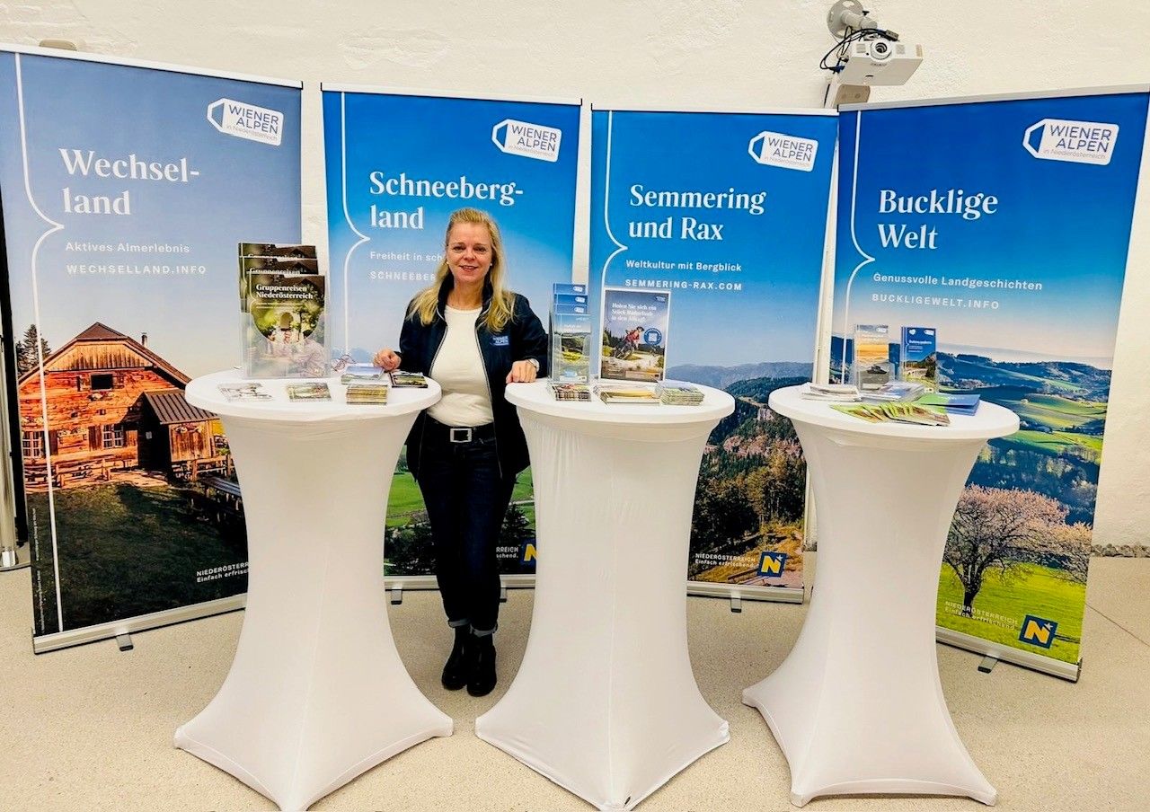 Vor vier Rückwänden einzelner Regionen der Wiener Alpen steht eine Frau hinter drei Stehtischen, auf denen verschiedene Broschüren angeboten werden. 